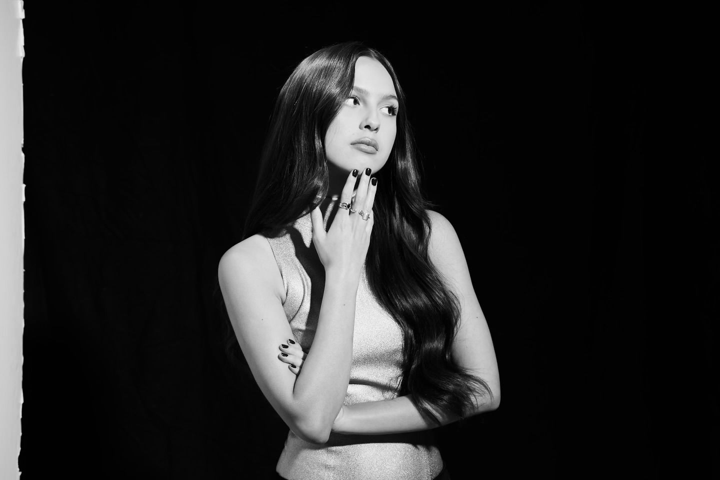 Singer-songwriter Olivia Rodrigo poses with her hand on her chin. Her body is set against an all-black background.